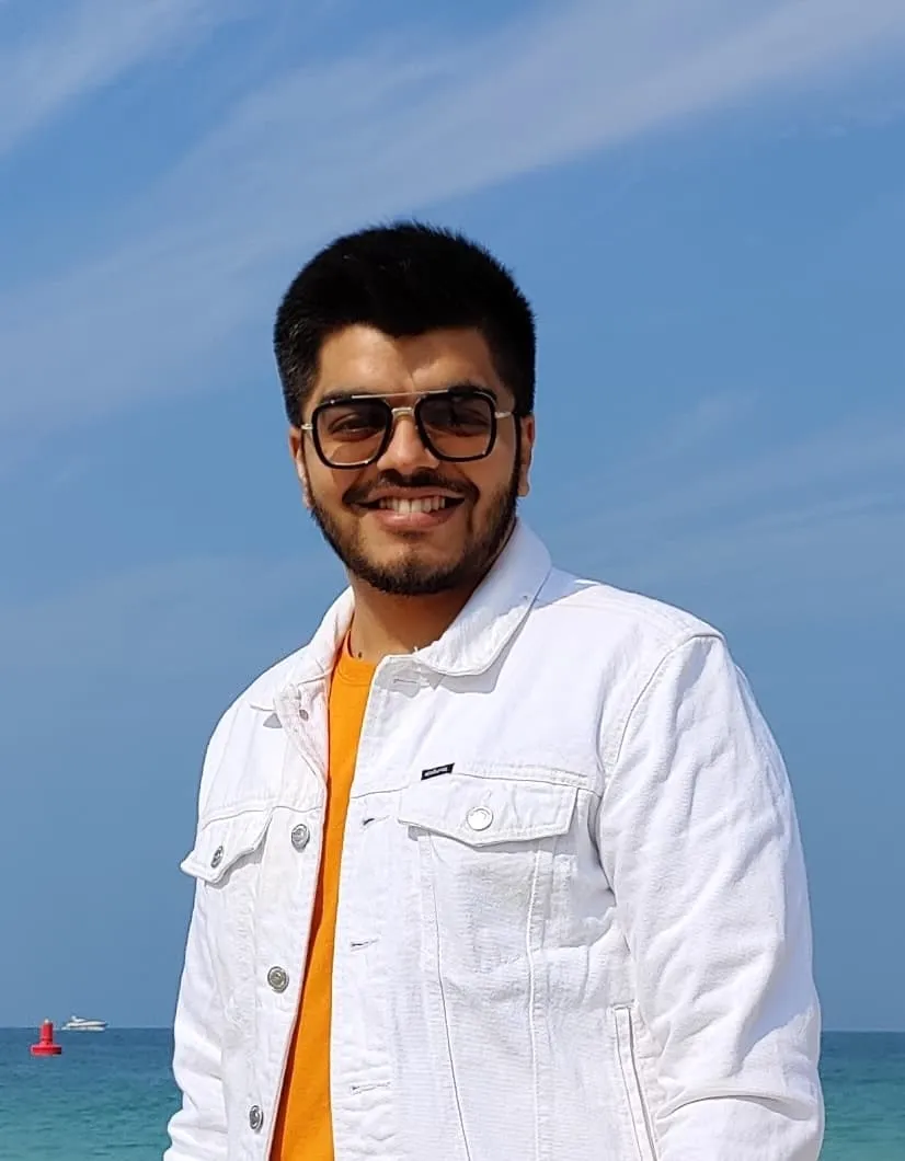 Shivam smiling in a white denim jacket by the sea